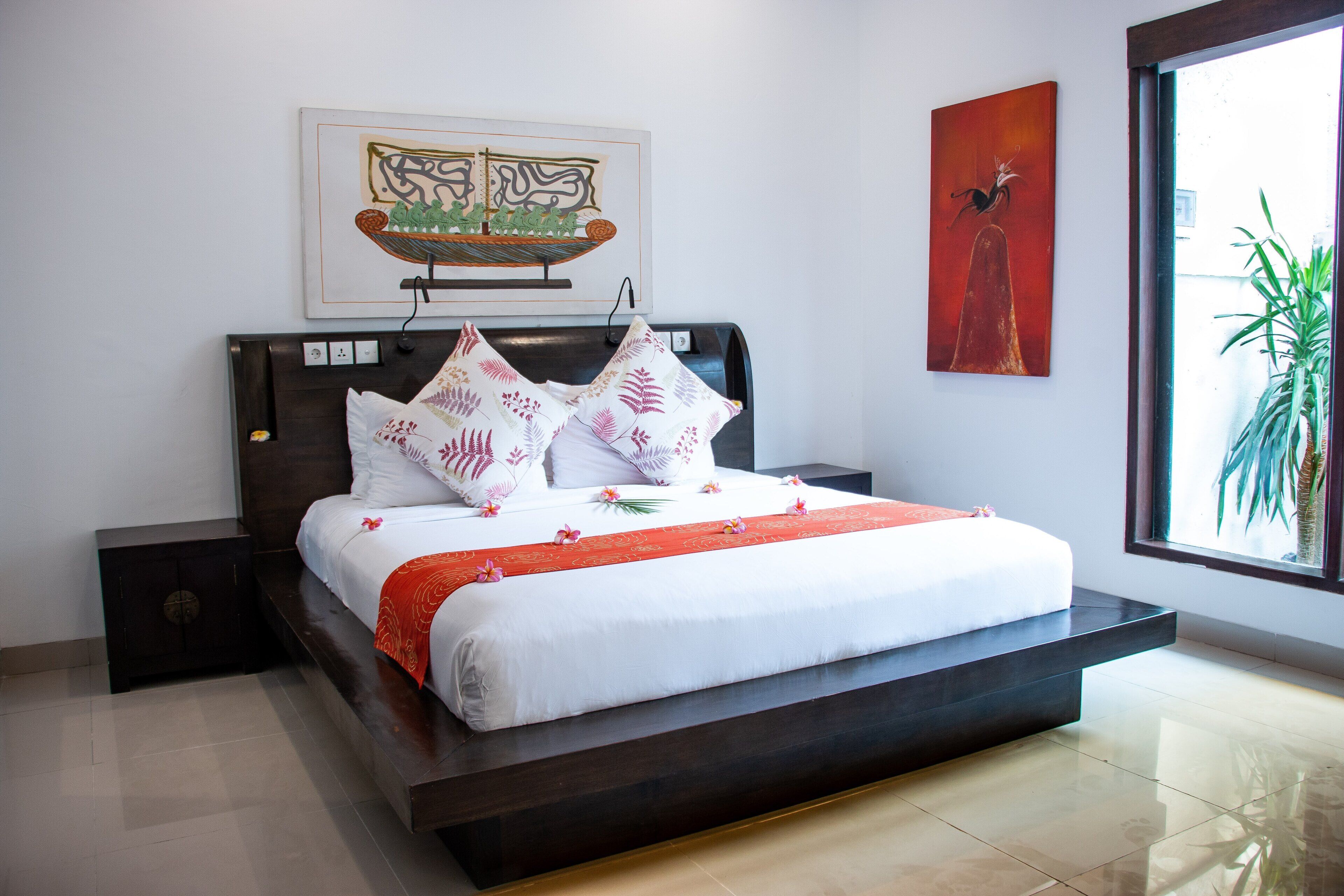 A modern bedroom with a large bed, a nightstand, a wall-mounted artwork, and a window with a plant.
