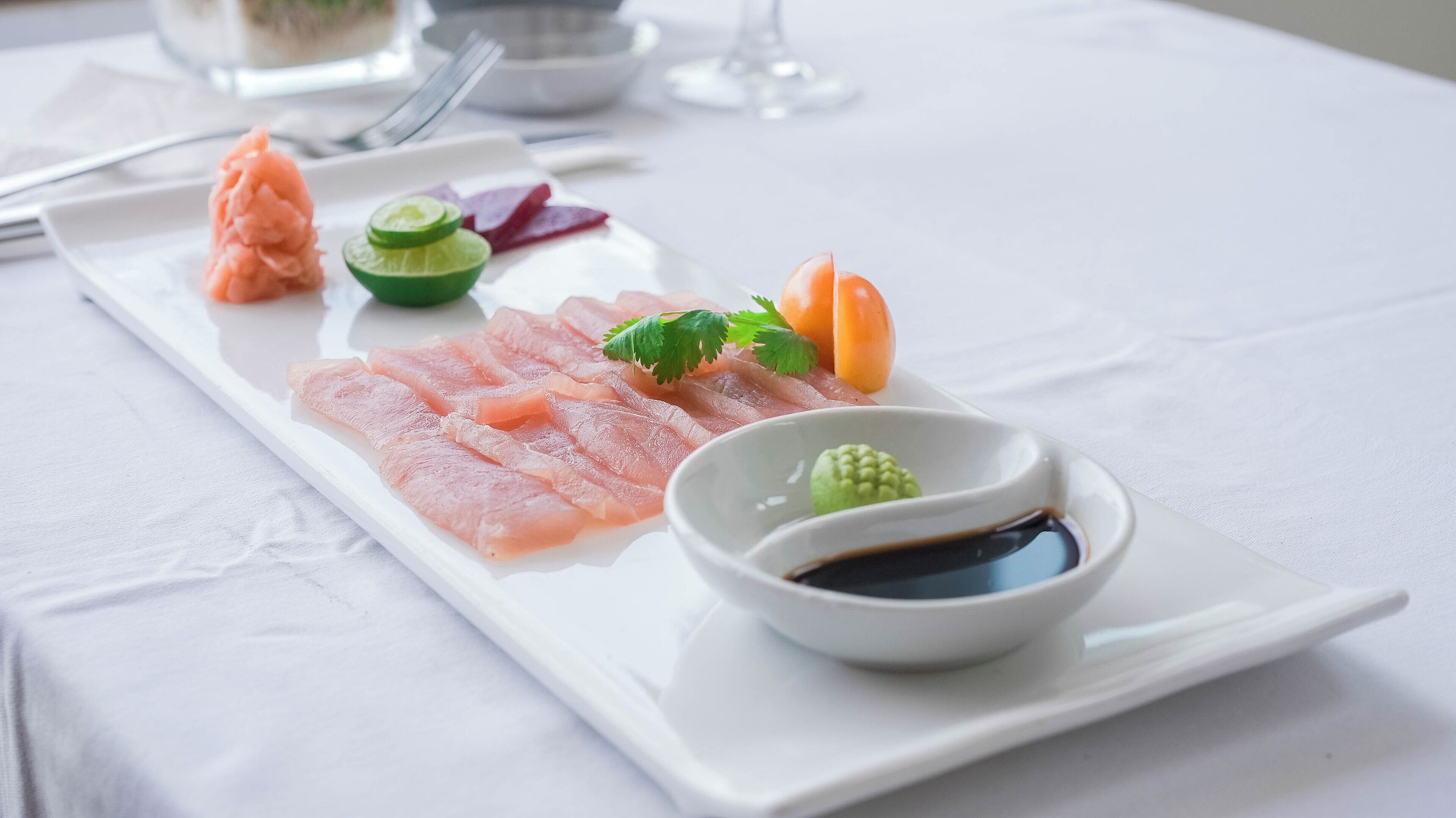A white rectangular plate with thinly sliced fish, garnishes, and a small bowl of sauce on a white tablecloth.