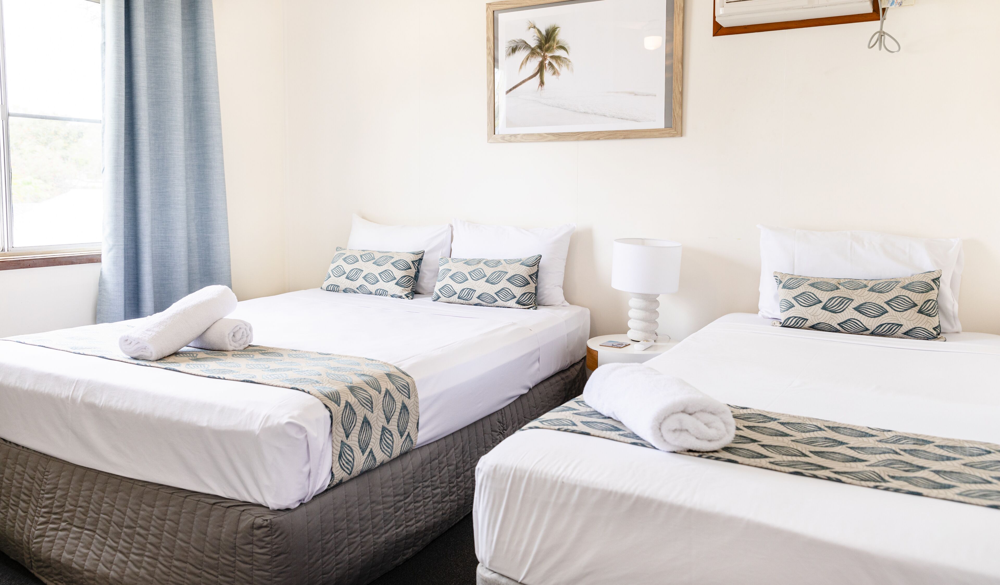 A hotel room with two beds, each with white linens and patterned pillows. There is a framed picture on the wall, a window with curtains, and a bedside table with a lamp.