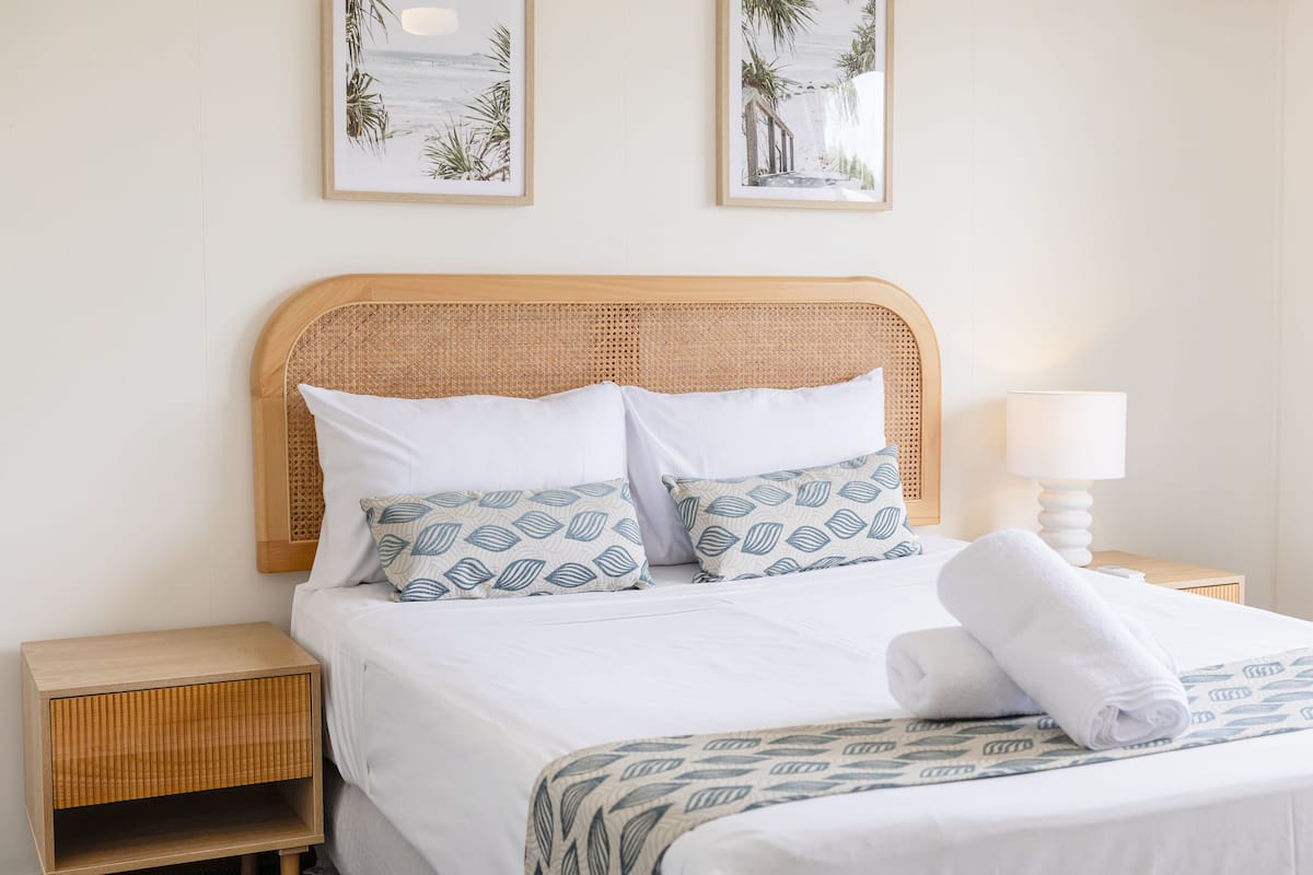 A neatly made bed with white linens and patterned pillows, a wooden nightstand with a lamp, and two framed pictures on the wall.