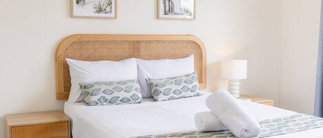 A neatly made bed with white linens and patterned pillows, a wooden nightstand with a lamp, and two framed pictures on the wall.