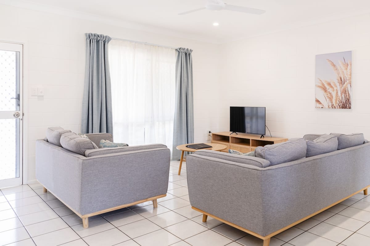 A modern living room with two grey sofas, a wooden TV stand, and a large window with curtains.