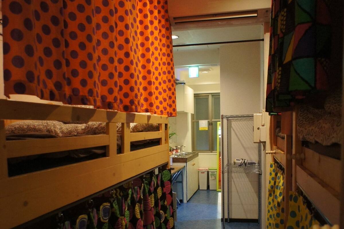 Un pasillo estrecho con una litera a la izquierda, una zona de cocina con refrigerador y lavabo en el centro, y una puerta que conduce a otra habitación a la derecha.