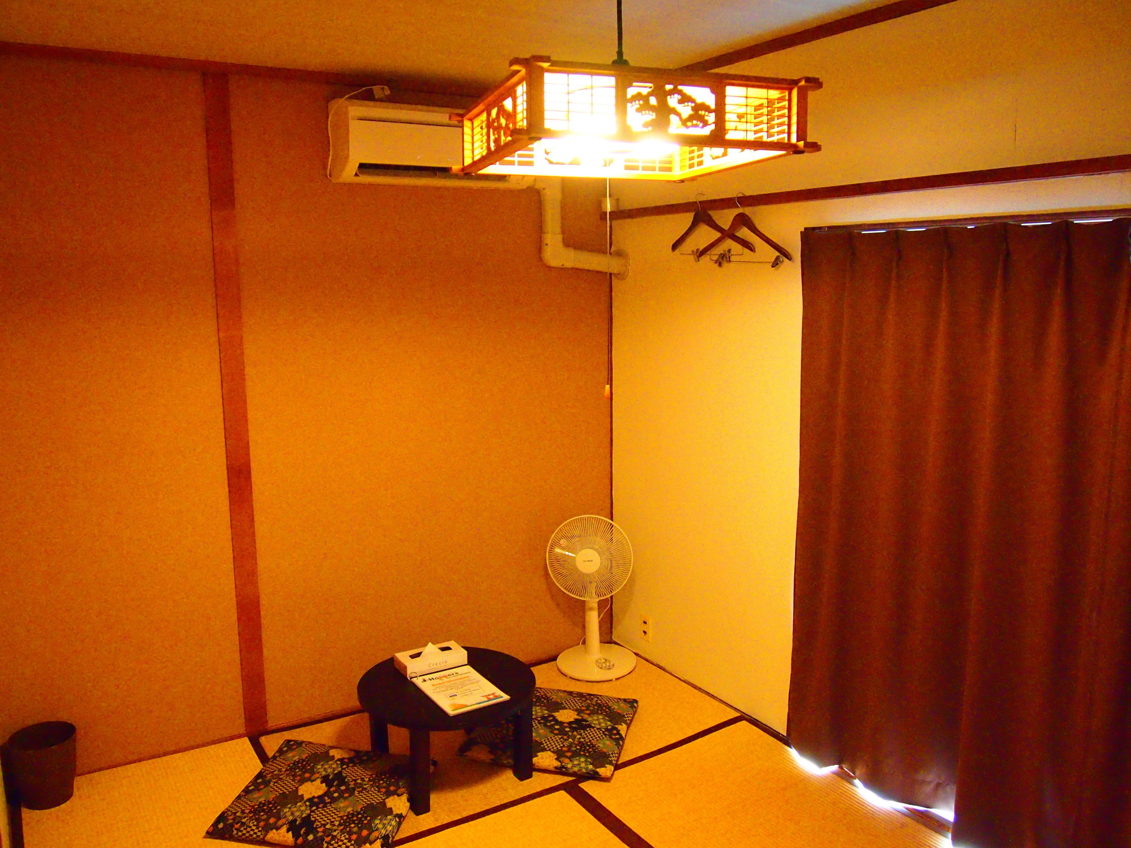 Una habitación con suelo de tatami, un ventilador de pie, una mesita con un libro y una puerta corredera tradicional japonesa.