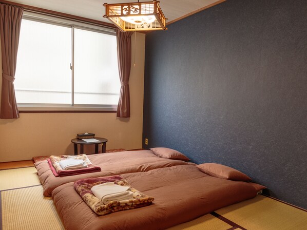 A traditional Japanese-style room with tatami flooring, a bed with pink bedding, a small table with a lamp, and a window with curtains.