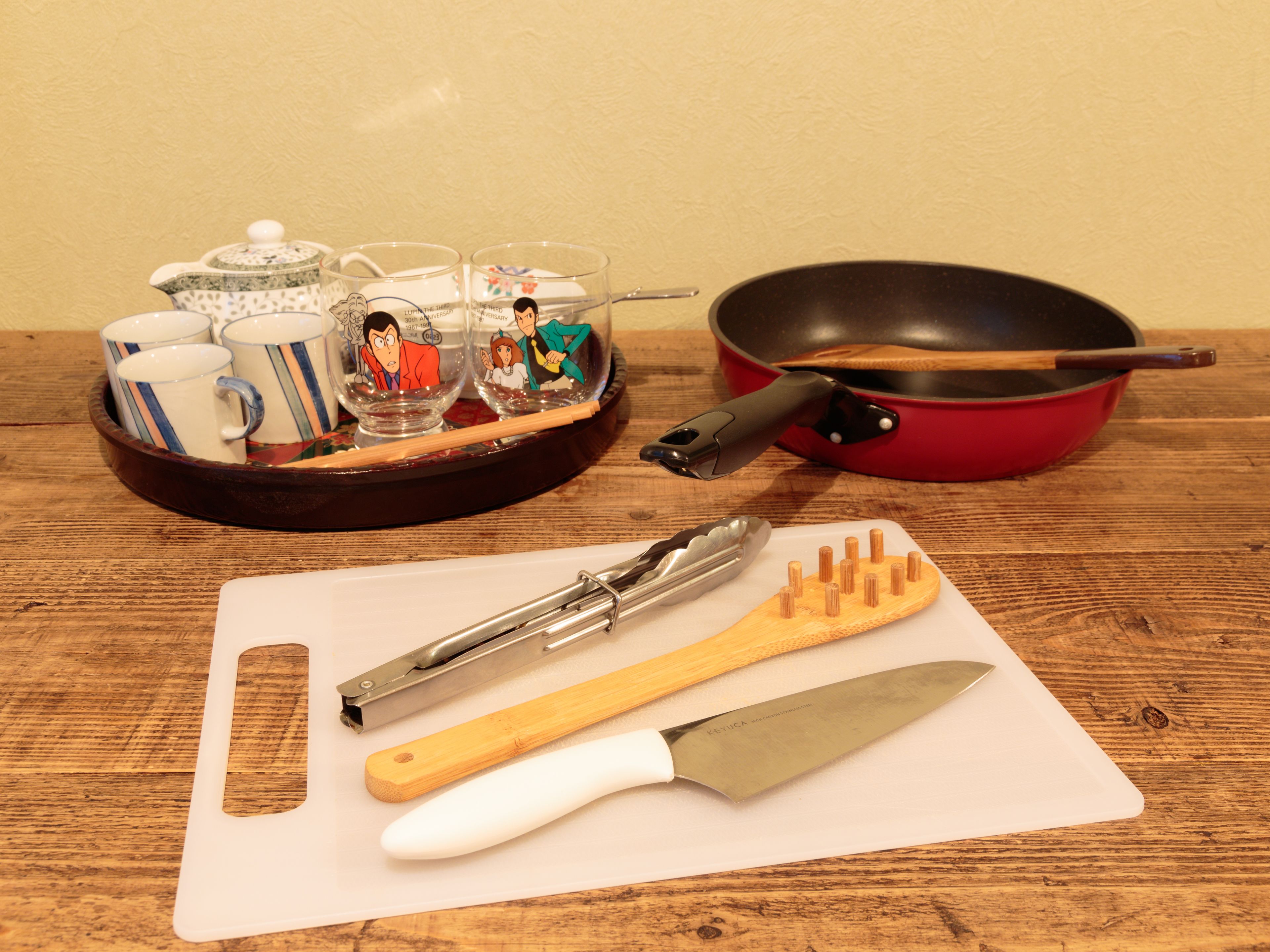 Una mesa de madera con una tabla de cortar, un cuchillo, unas pinzas, una sartén y una bandeja con tazas y adornos.