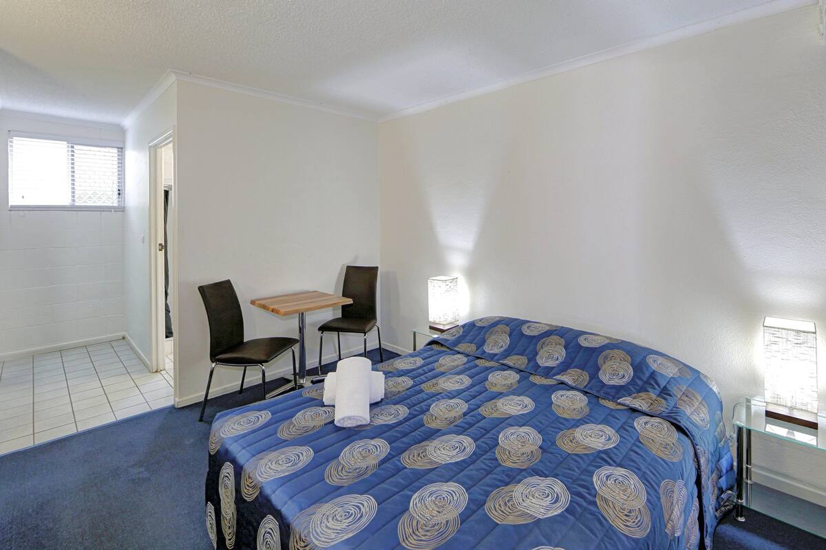 Quarto de hotel com uma cama grande, uma mesinha, duas cadeiras, uma janela e uma porta.