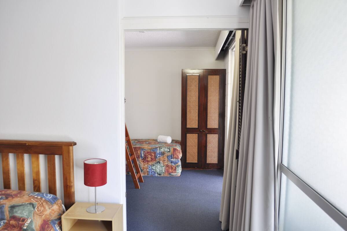 Quarto de hotel com uma cama, uma mesinha com abajur, uma mala e uma porta que dá para outro cômodo.
