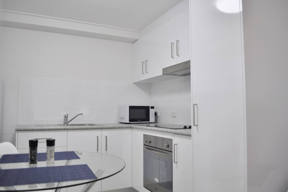 Uma cozinha moderna com armários brancos, forno de aço inoxidável, micro-ondas e mesa de vidro com um jogo americano azul.