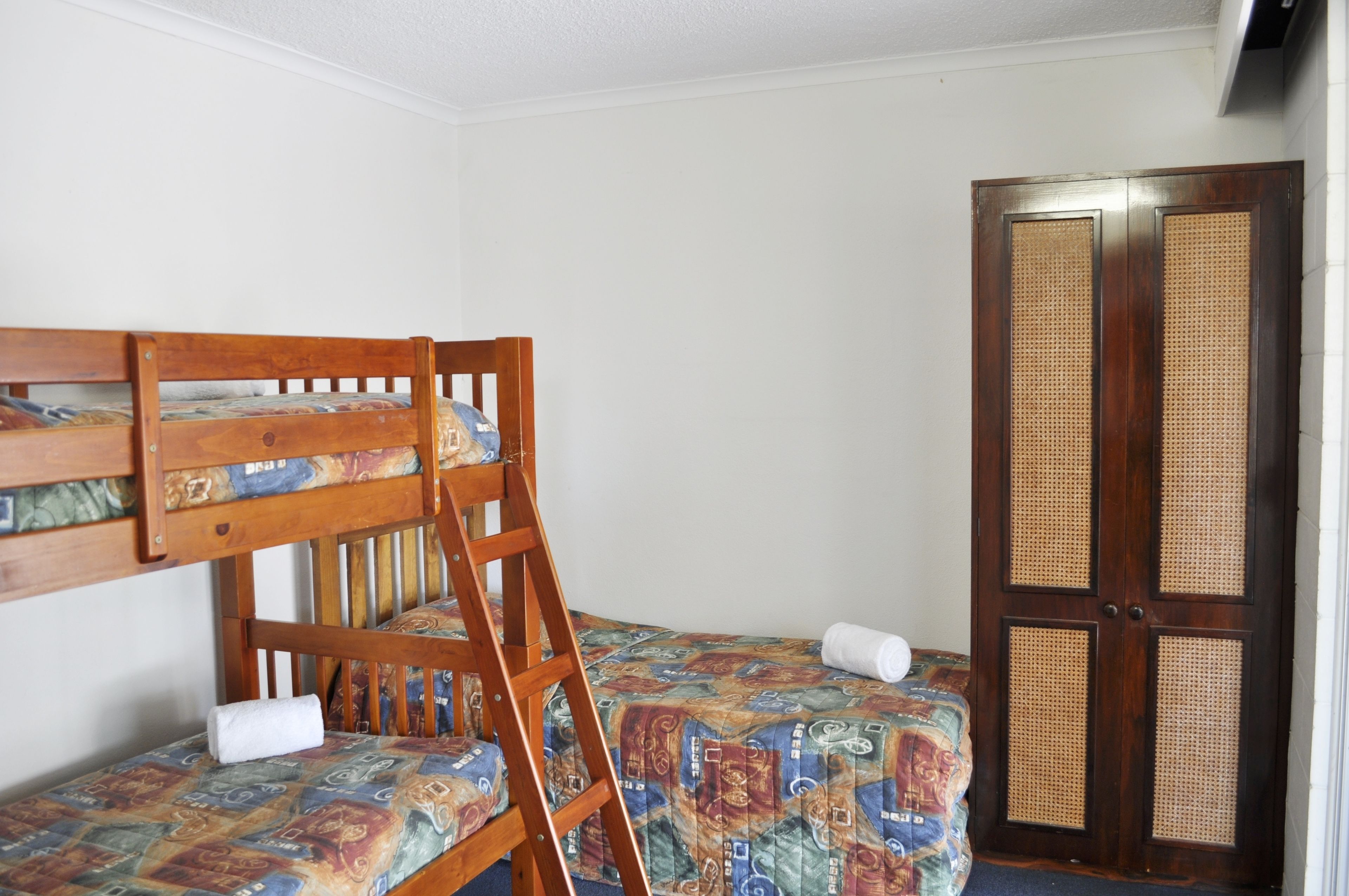 A room with a bunk bed, a wooden ladder, a door with a woven panel, and a patterned bedspread.