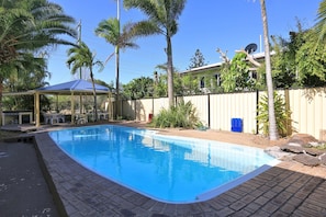 A rectangular swimming pool with a tiled edge, surrounded by a brick patio. There is a covered seating area with chairs and a table to the left of the pool. - Alexandra Park Motor Inn (Bundaberg West)