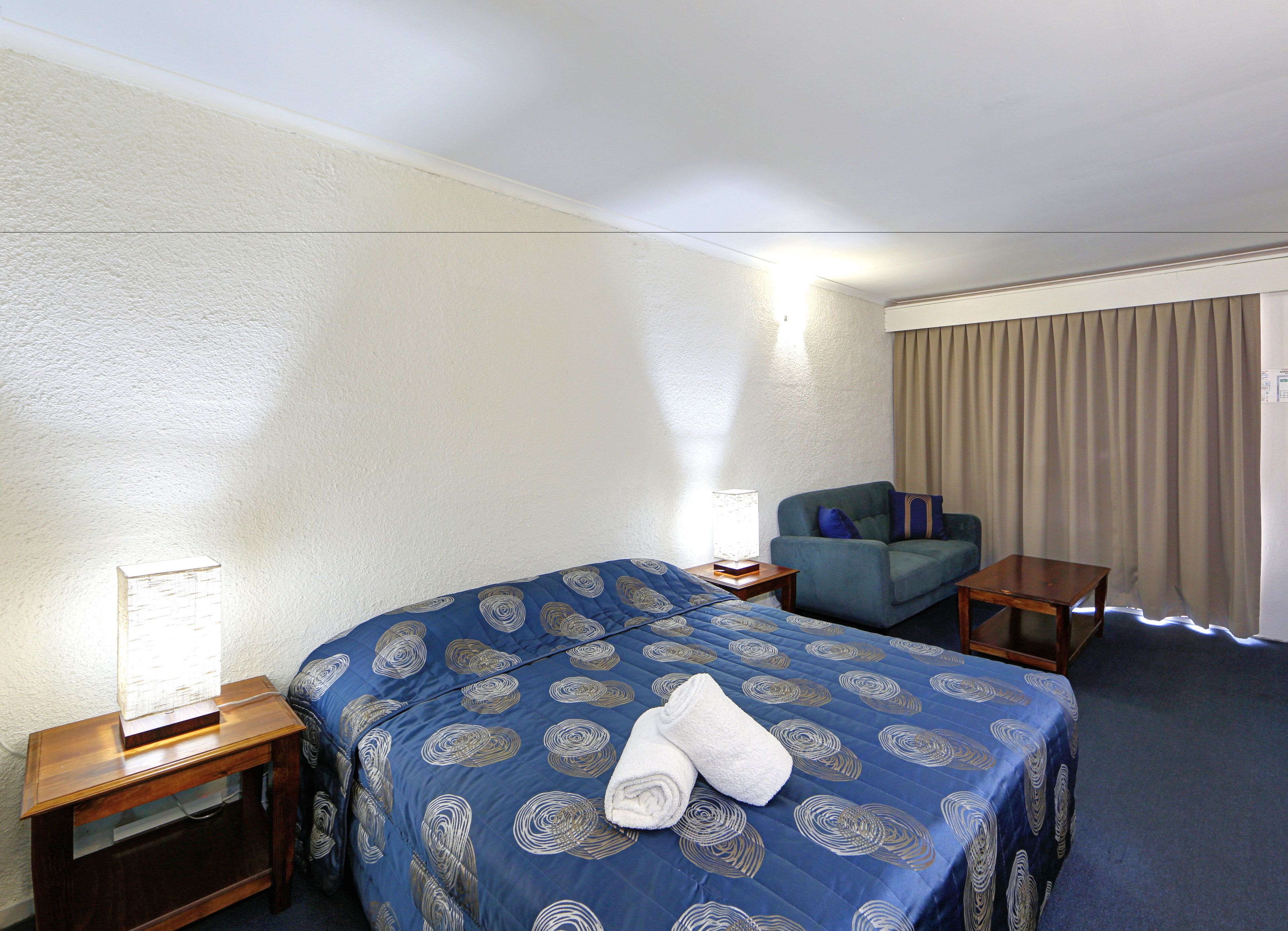 A hotel room with a bed, a chair, a small table, a lamp, and a window with curtains.