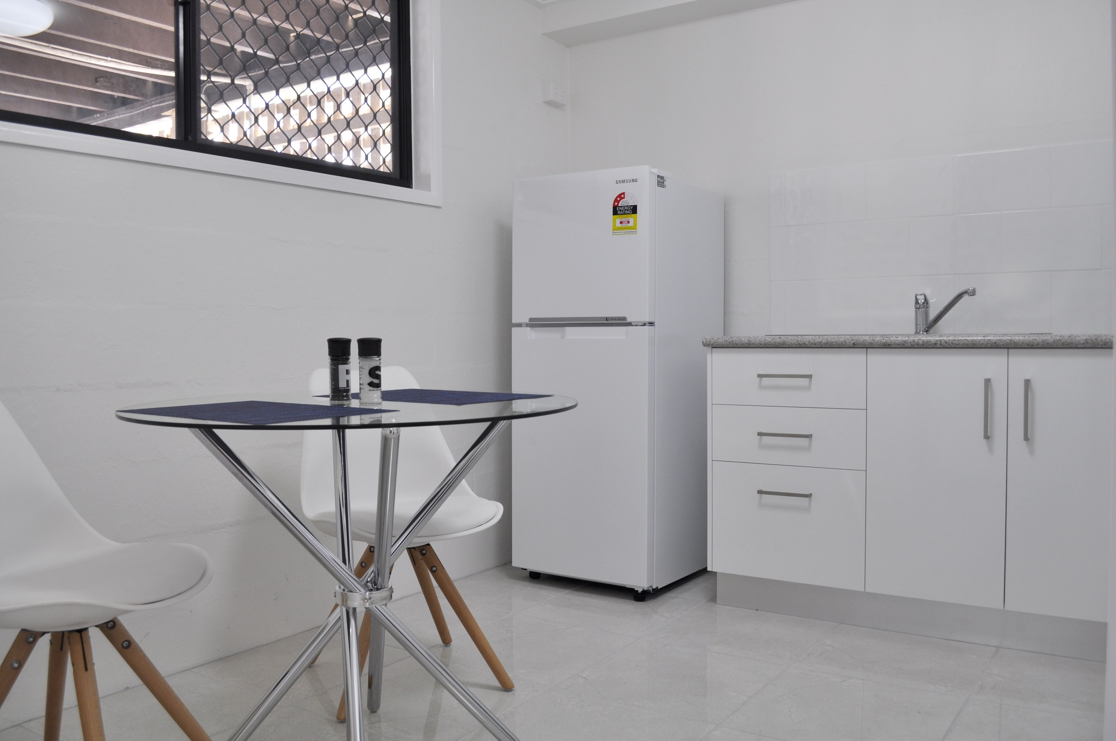 A compact kitchen with a white refrigerator, a small sink, and a glass-top table with chairs.