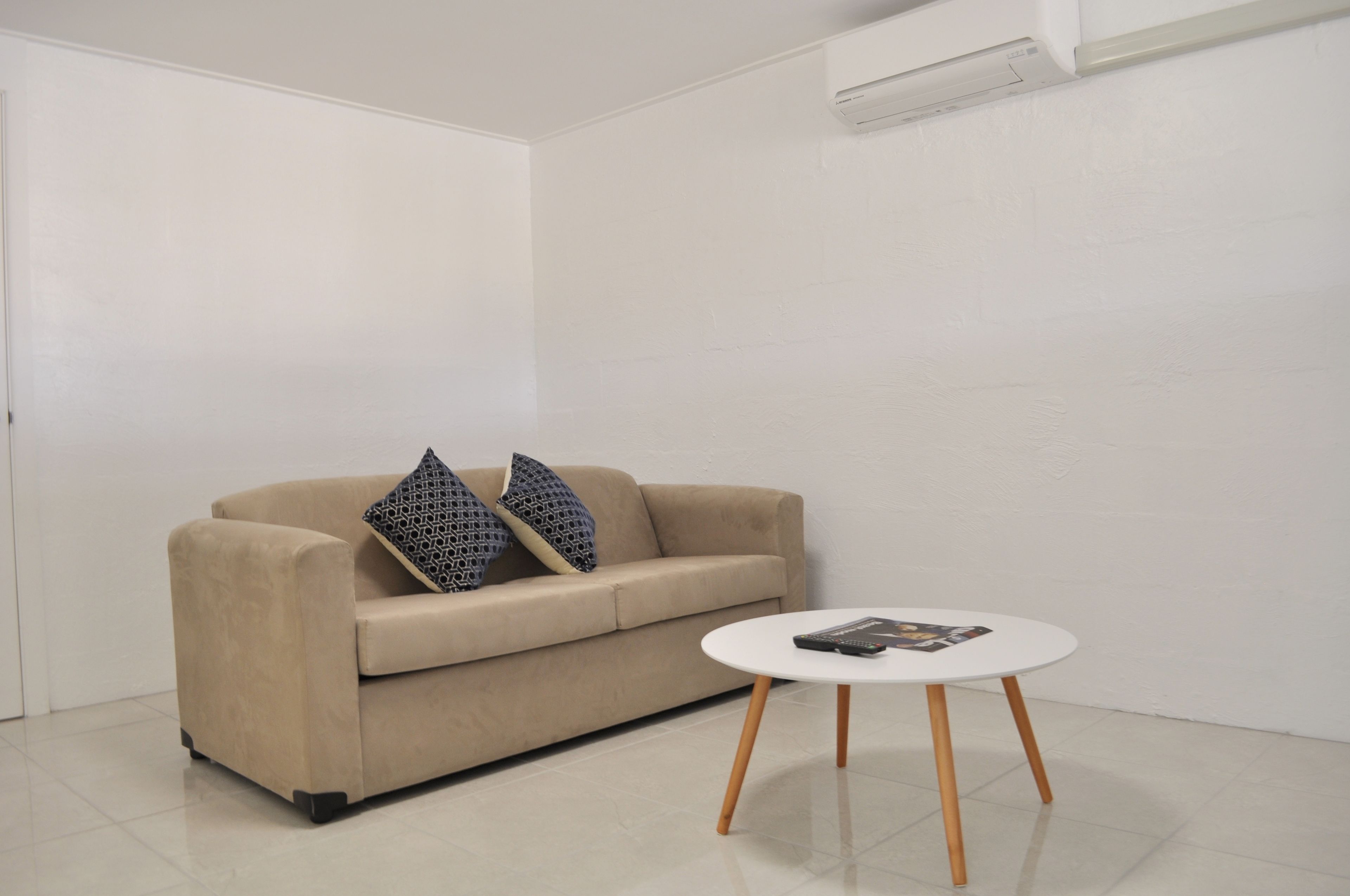 Un salón minimalista con un sofá color beige, dos almohadones estampados y una pequeña mesa de centro blanca con un control remoto y una revista.