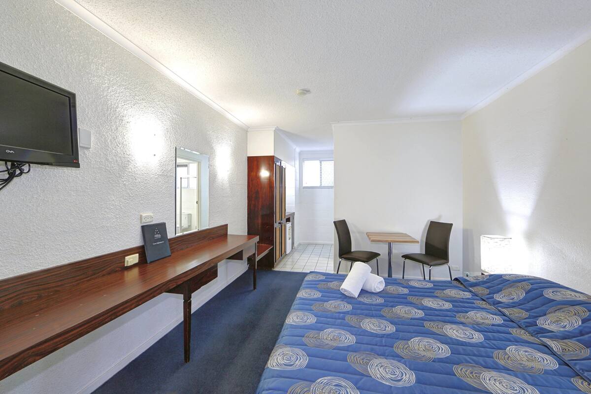 Quarto de hotel com cama, escrivaninha, cadeira, televisão e janela.