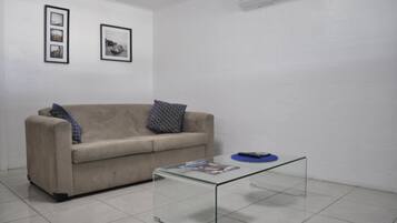 Een minimalistisch ingerichte kamer met een beige bank, een glazen salontafel en twee ingelijste foto's aan de muur.