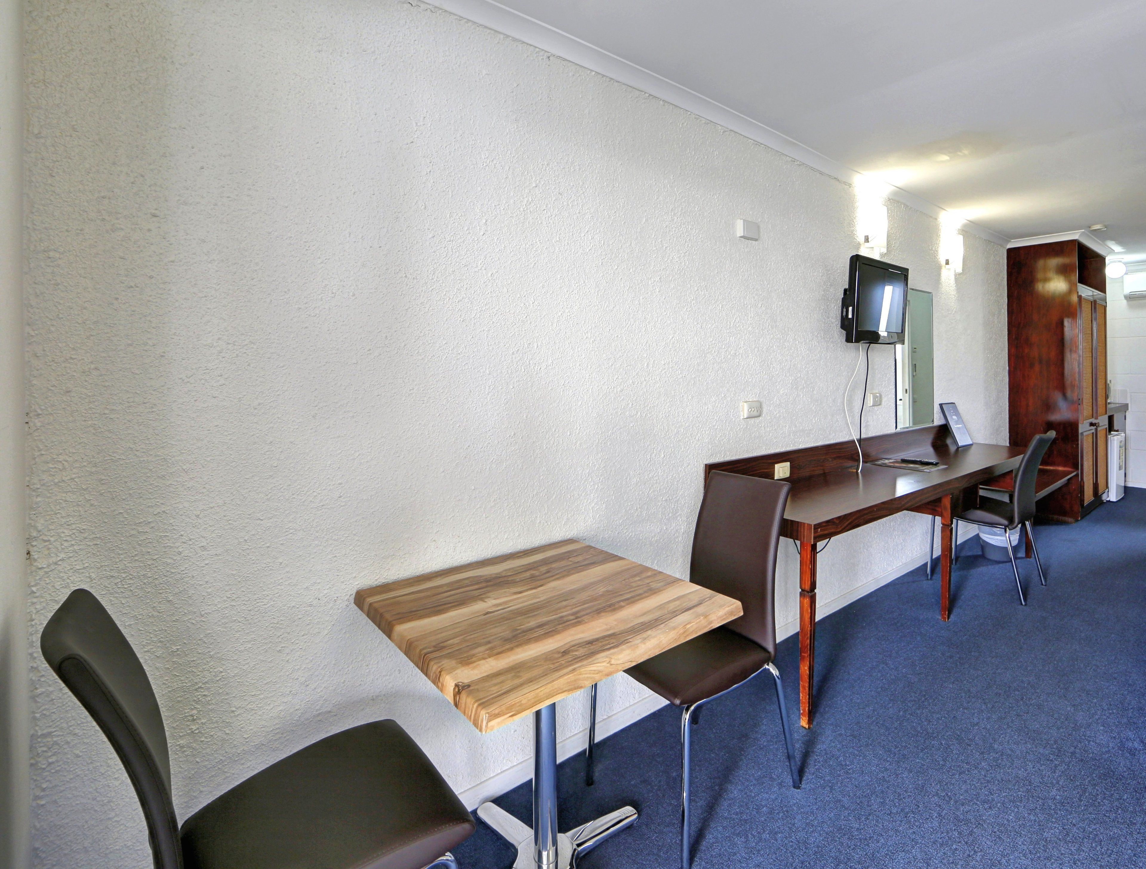 A hotel room with a wooden desk, two chairs, a television mounted on the wall, and a door leading to another room.