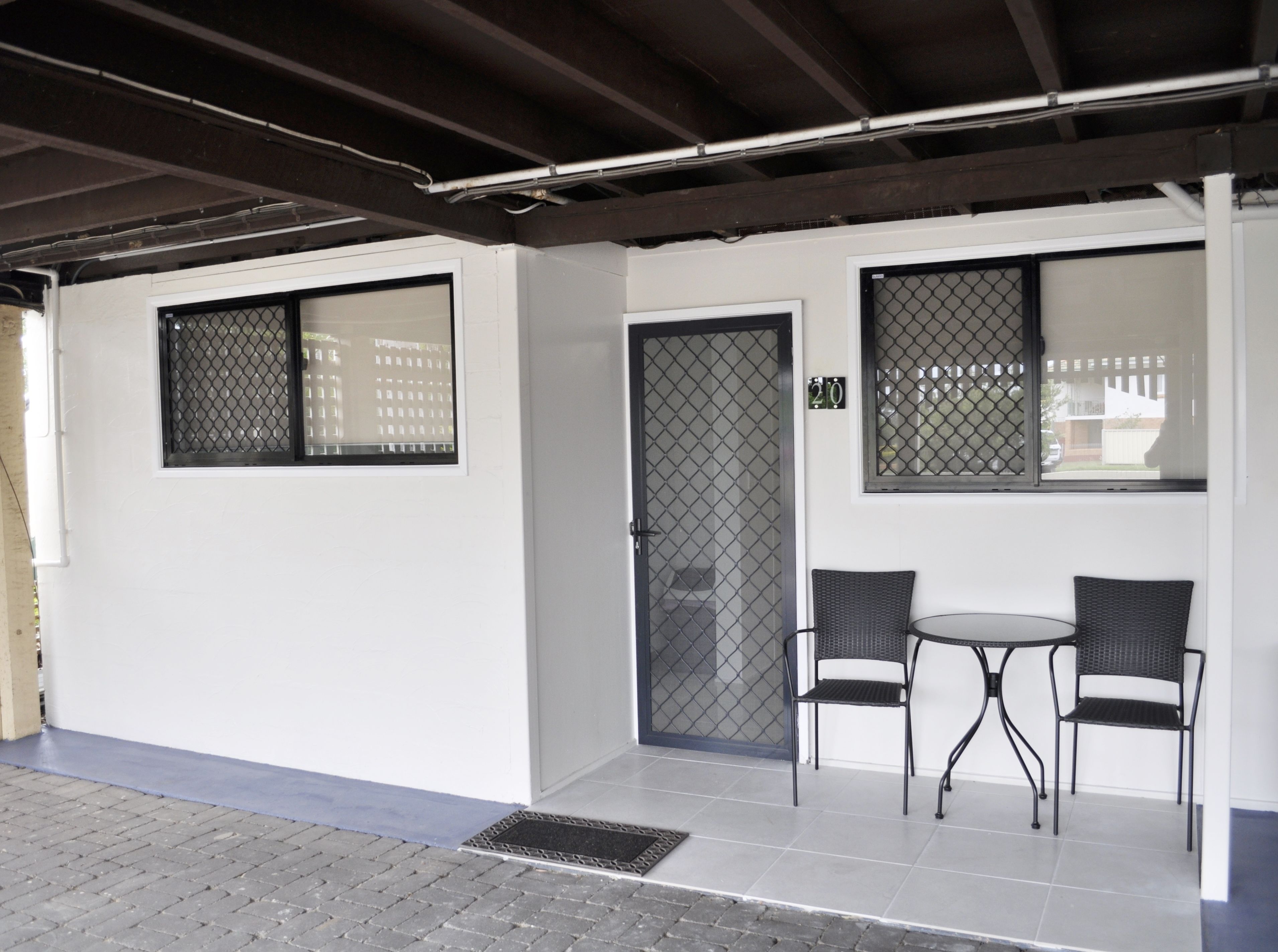 A covered outdoor seating area with two chairs and a table, a door with a security screen, and a window with a view of buildings.
