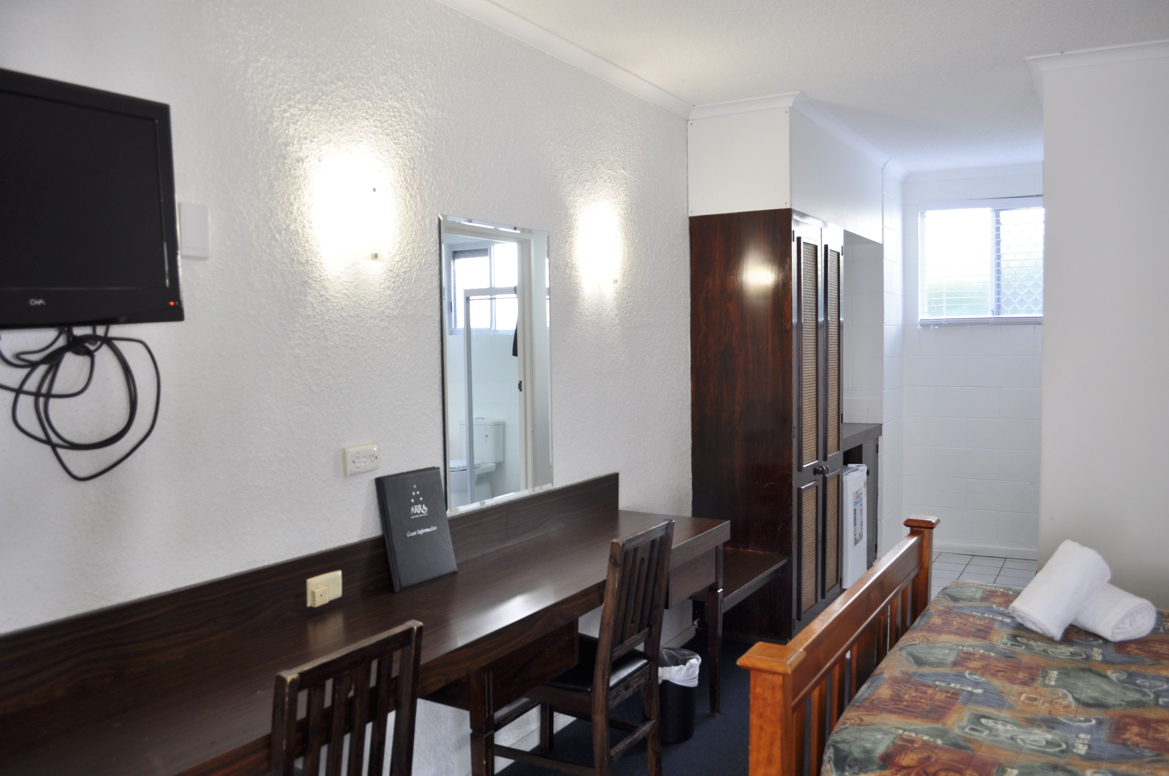 A hotel room with a wooden desk, a chair, a bed with a colorful bedspread, a TV mounted on the wall, a mirror, and a window with blinds.