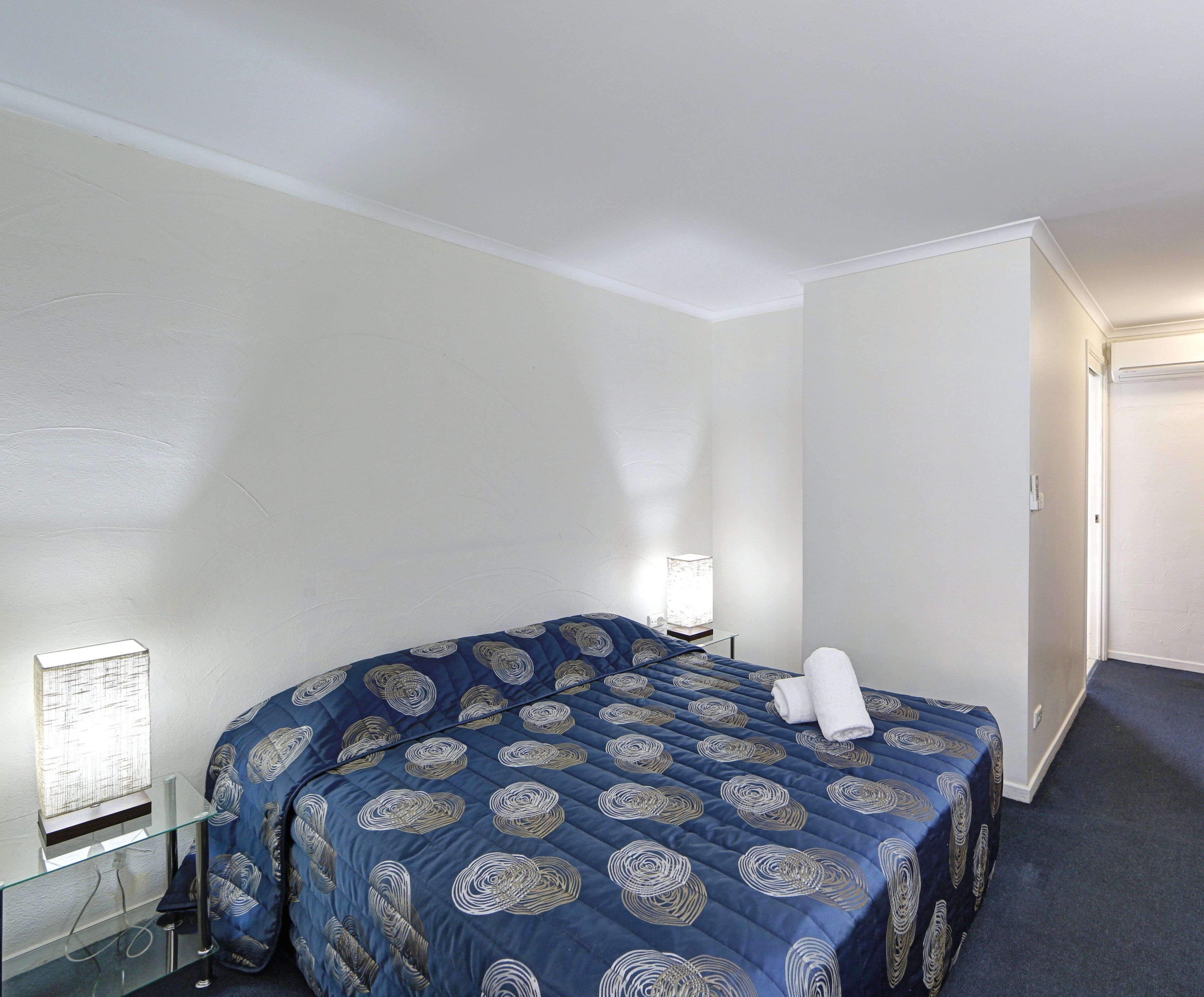 A hotel room with a bed, a bedside table, a lamp, and a window with blinds.