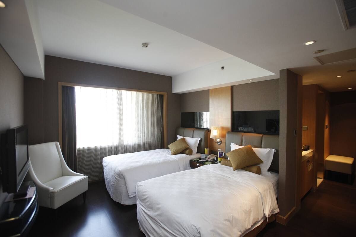 A hotel room with two beds, a TV, a desk, and a window with curtains.