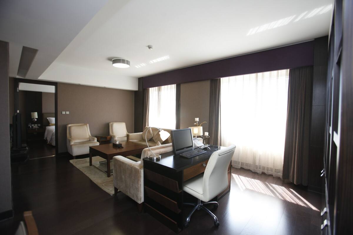 A modern hotel room with a desk, two chairs, a sofa, and a large window with curtains.