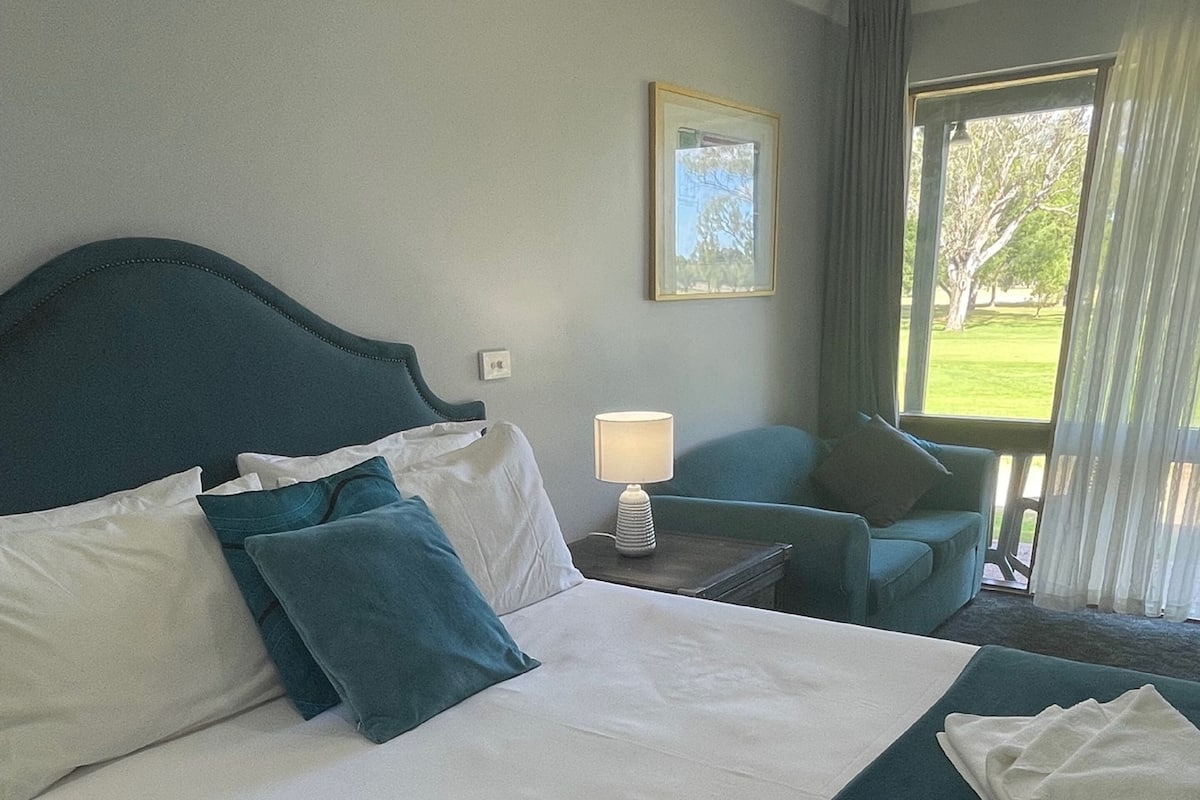 A neatly made bed with white and teal bedding, a bedside lamp, and a view of a green landscape through a window.