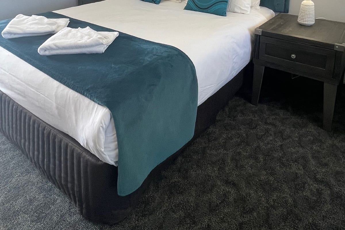 A neatly made bed with a teal bedspread and white linens, two bedside tables with lamps, and a dark carpeted floor.