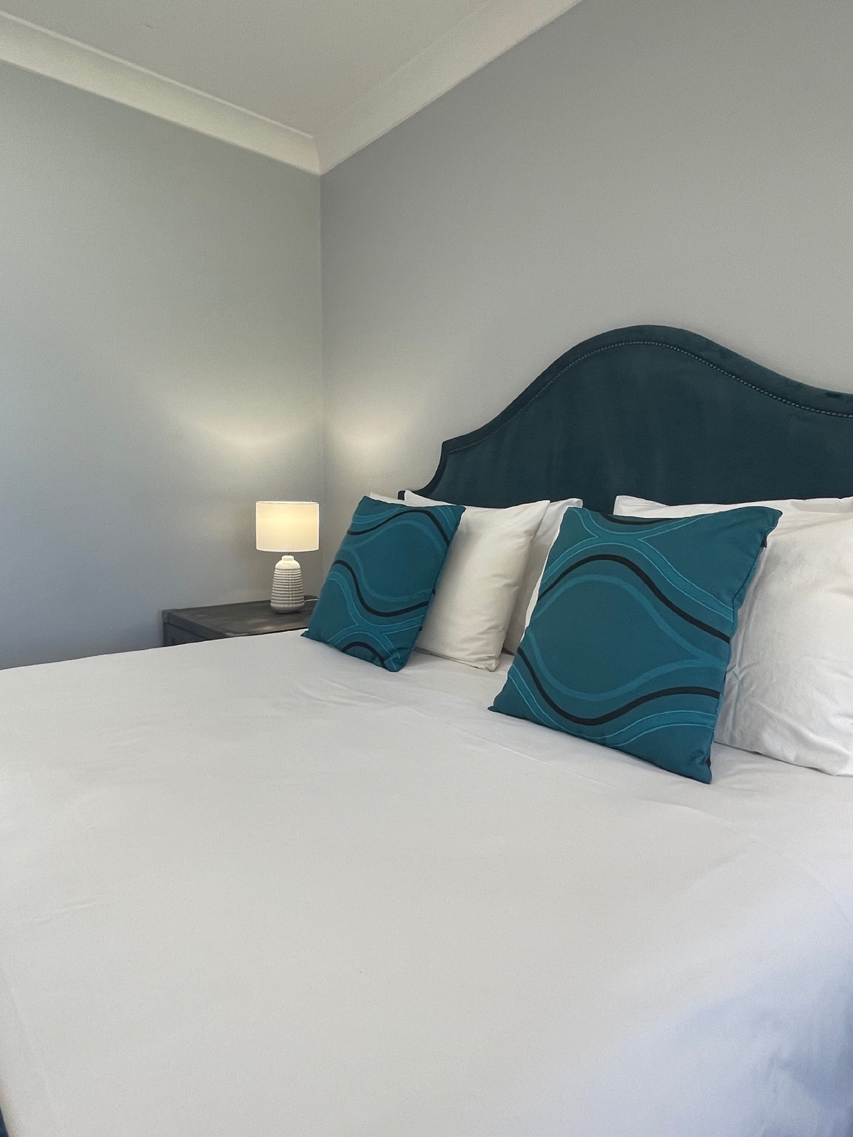 Un lit bien fait, avec du linge de lit blanc et des coussins à motifs turquoise, une lampe de chevet et une tête de lit sombre, le tout sur un mur de couleur claire.