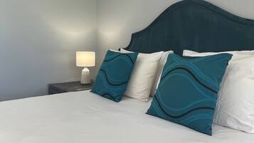 Un lit bien fait, avec du linge de lit blanc et des coussins à motifs turquoise, une lampe de chevet et une tête de lit sombre, le tout sur un mur de couleur claire.