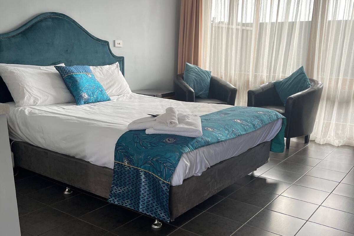 A neatly made bed with a teal headboard, white bedding, and a blue patterned throw. There are two chairs with teal cushions and a window with sheer curtains.