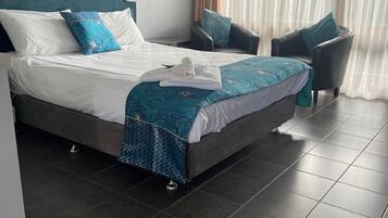 Una cama bien hecha con un cabecero verde azulado, ropa de cama blanca y una manta estampada azul. Hay dos sillas con cojines verde azulado y una ventana con cortinas de encaje.
