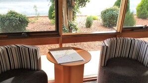 Une pièce avec deux fauteuils à rayures, une petite table ronde sur laquelle est posée une carte, et une vue sur un jardin aménagé, visible à travers de grandes fenêtres.