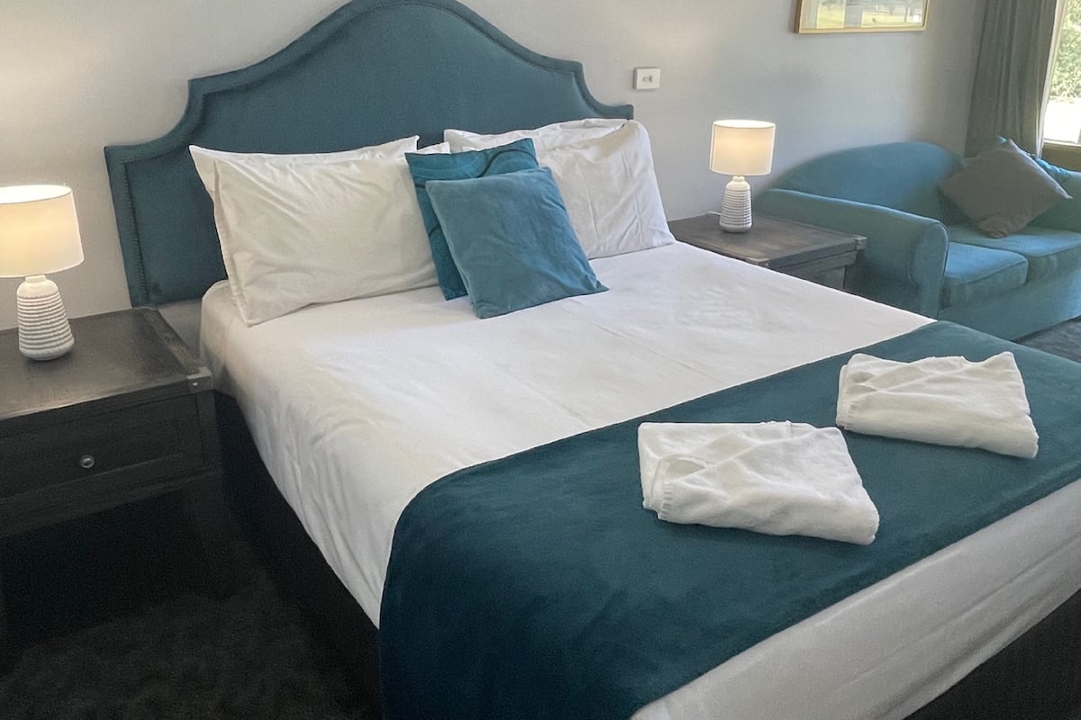 A hotel room with a large bed, two bedside tables, a nightstand, a blue armchair, and a window with curtains.