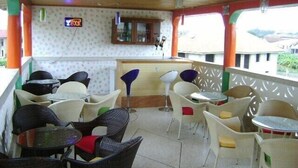 Un espace extérieur aménagé, abrité et doté de chaises et de tables en osier, un coin bar avec un panneau lumineux au néon, et une vue sur le paysage environnant.