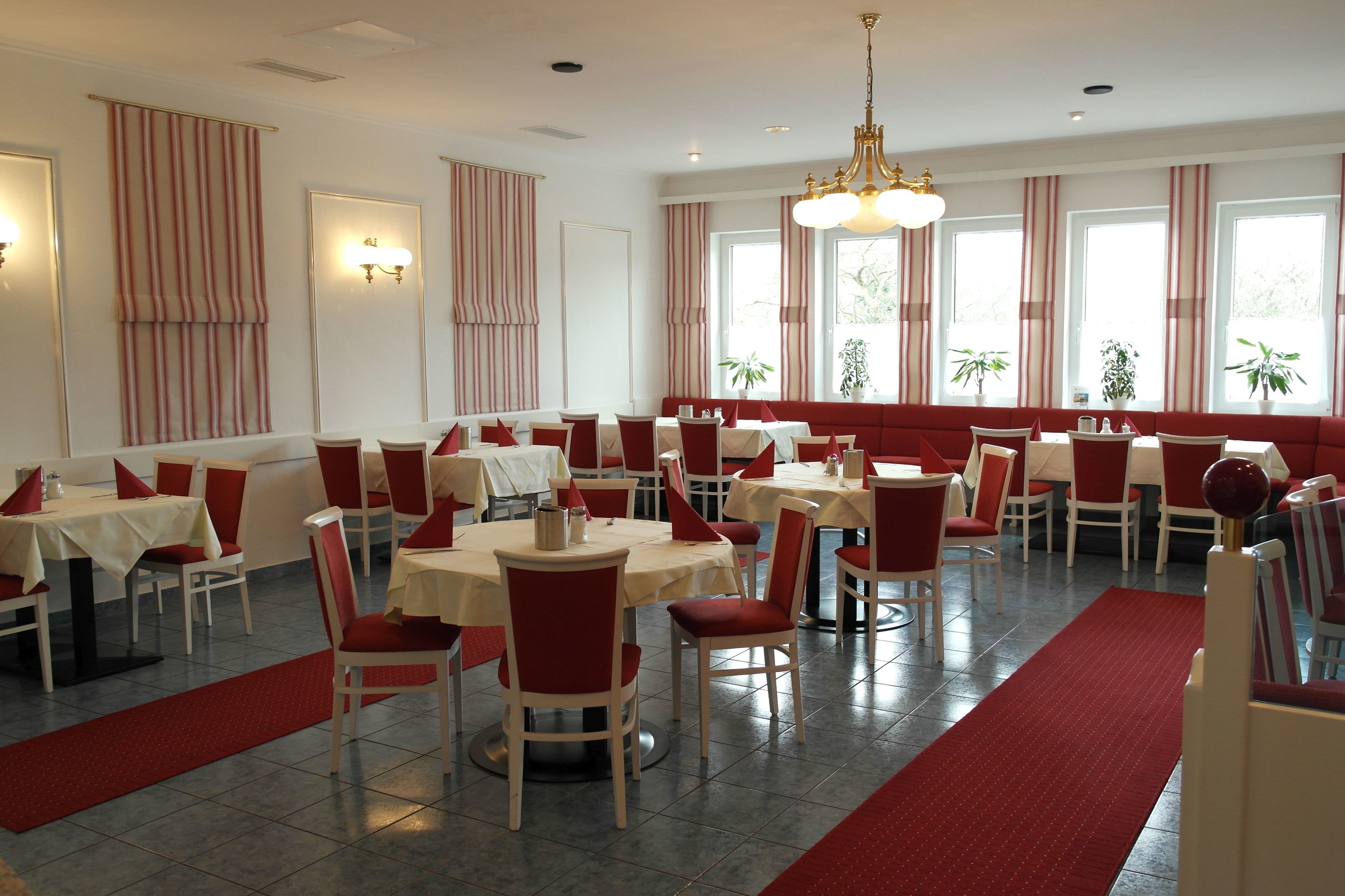 Un comedor con mesas y sillas, alfombra roja y ventanas amplias con cortinas.