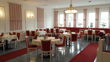 Un comedor con mesas y sillas, alfombra roja y ventanas amplias con cortinas.