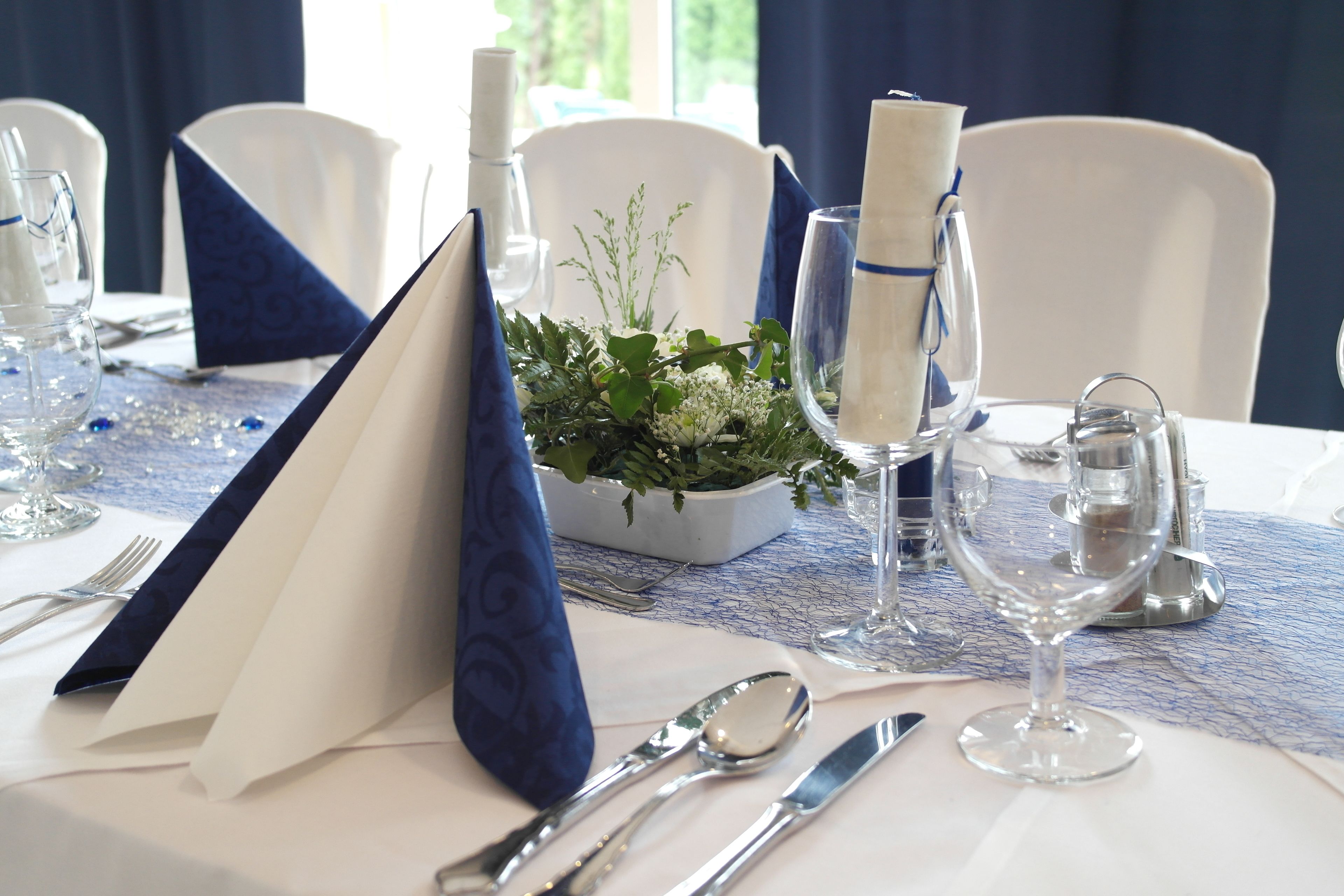 Una mesa dispuesta para comer, con mantel azul, servilletas blancas y un centro de mesa con follaje.