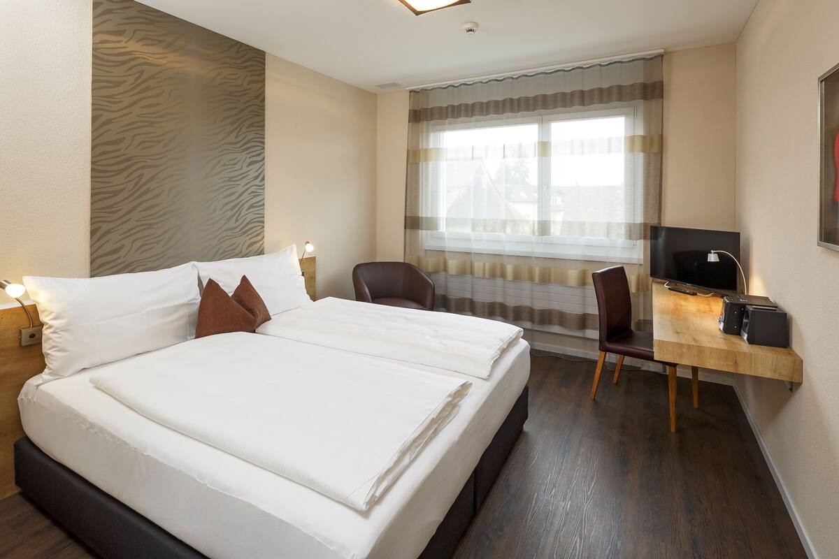 Ein Hotelzimmer mit einem Bett, einem Schreibtisch mit Computer, einem Stuhl und einem Fernseher.