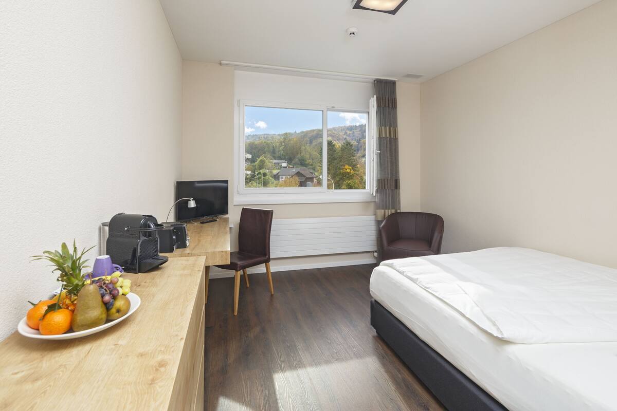 Ein Hotelzimmer mit Bett, Schreibtisch, Computer, Stuhl und Blick ins Freie.