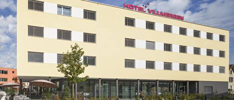 Un hotel con fachada amarilla, numerosas ventanas y un cartel que dice "HOTEL VILLMERGEN".