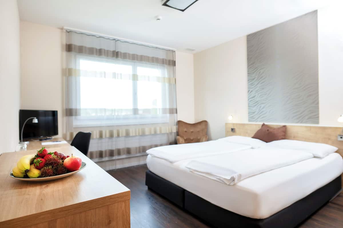 Ein Hotelzimmer mit einem Bett, einem Schreibtisch mit Obst, einem Sessel, einem Fernseher und einem Fenster mit Vorhängen.