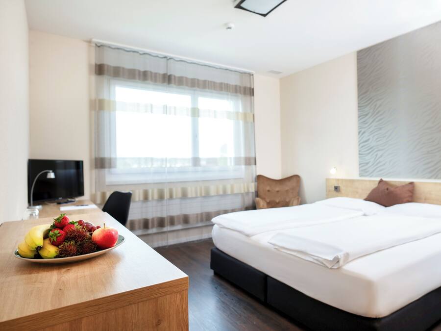 A hotel room with a bed, a desk with fruit, a chair, a TV, and a window with curtains.