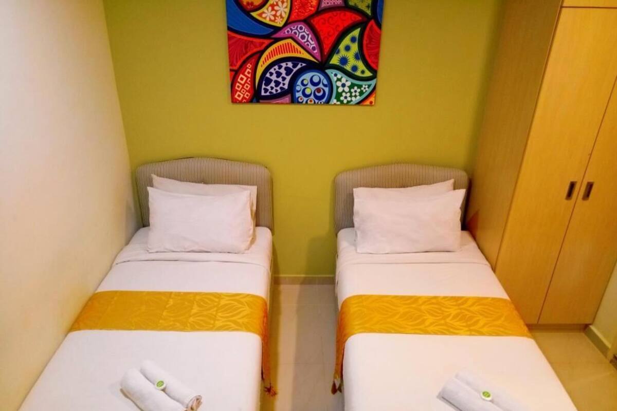 Two twin beds with white linens and yellow sashes, a colorful abstract painting on the wall, and a wooden wardrobe.
