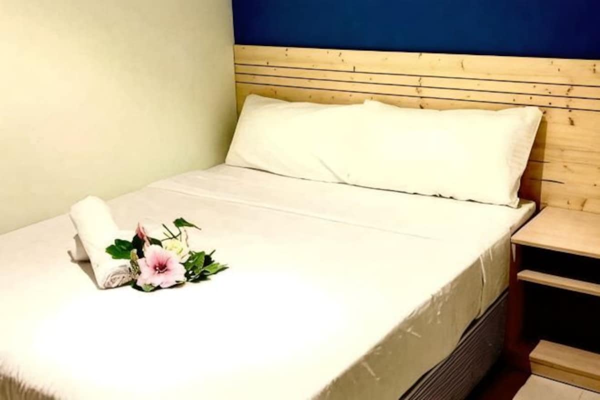Una cama bien hecha con sábana blanca, una toalla enrollada y un pequeño arreglo floral.