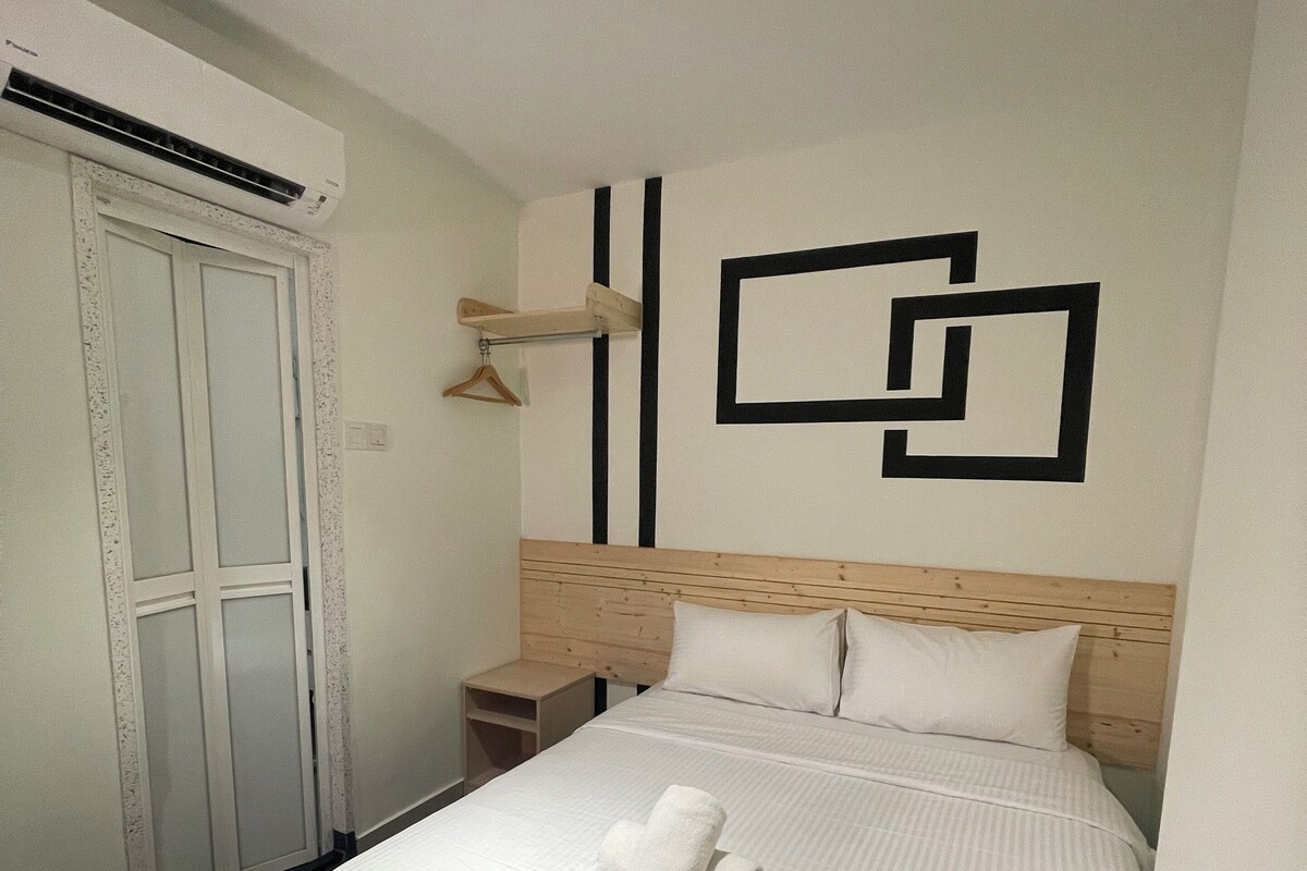Ein Hotelzimmer mit einem ordentlich bezogenem Bett, einem Holzkopfteil, einer Wand mit geometrischen Mustern, einer hängenden Leuchte und einer Tür mit satinierten Glasscheiben.