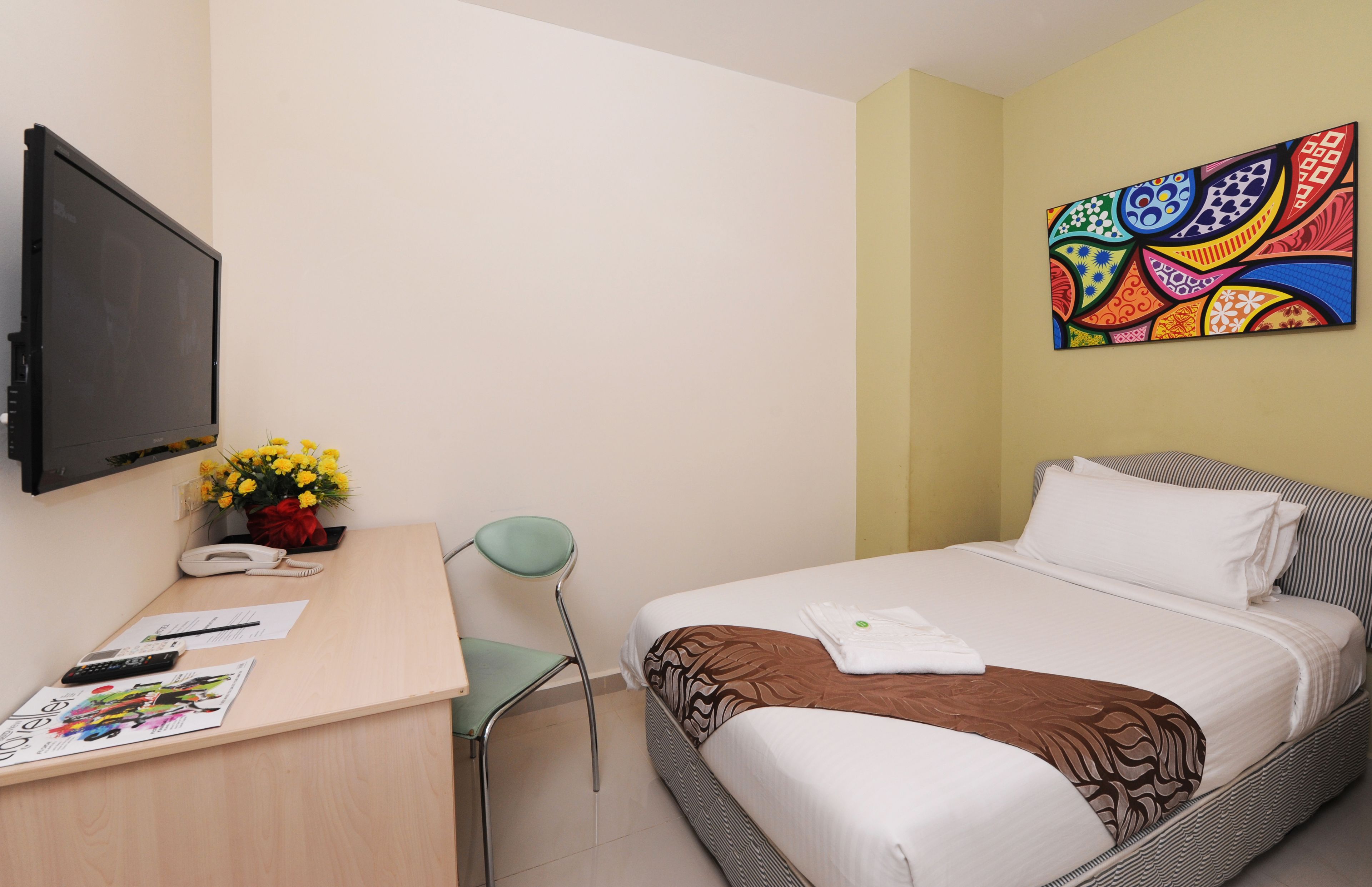 A hotel room with a bed, a desk with a phone, a chair, a TV, and a colorful wall art piece.