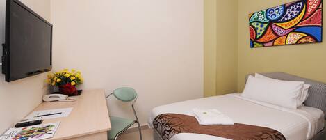 A hotel room with a bed, a desk with a phone, a chair, a TV, and a colorful wall art piece.