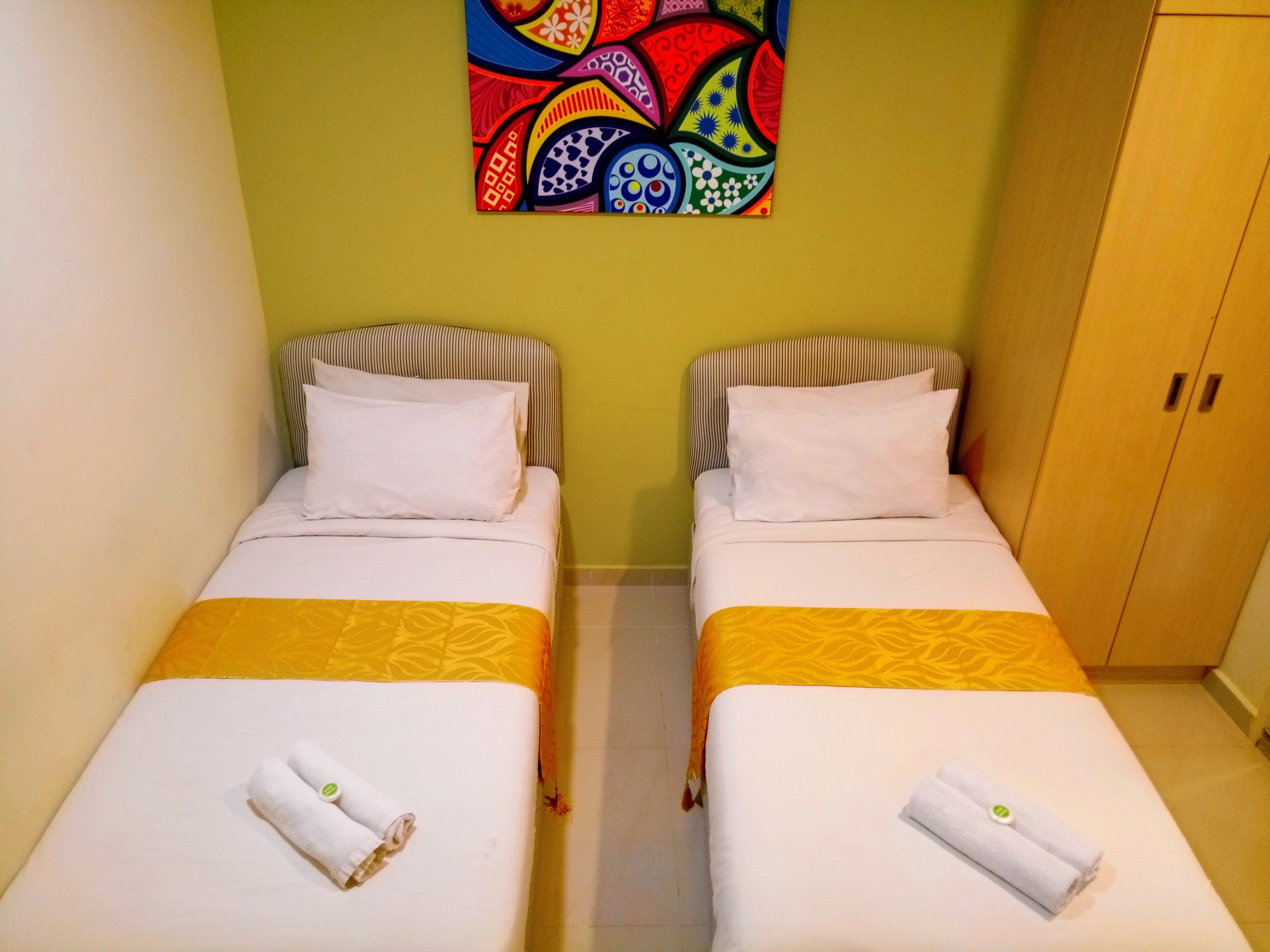 A twin room with two single beds, each with a white pillow and a rolled towel. The room has a green wall and a colorful abstract painting above the beds.