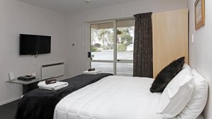 A hotel room with a bed, a flat-screen TV mounted on the wall, a bedside table with a lamp, and a view of the outdoors through a sliding glass door.